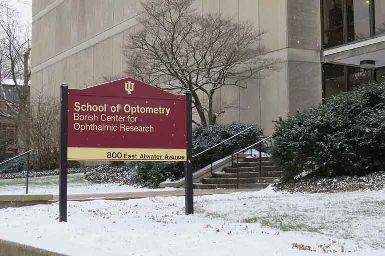 An image of the sign of the School of Optometry with the school in the background on a wintery day with snow on the ground.